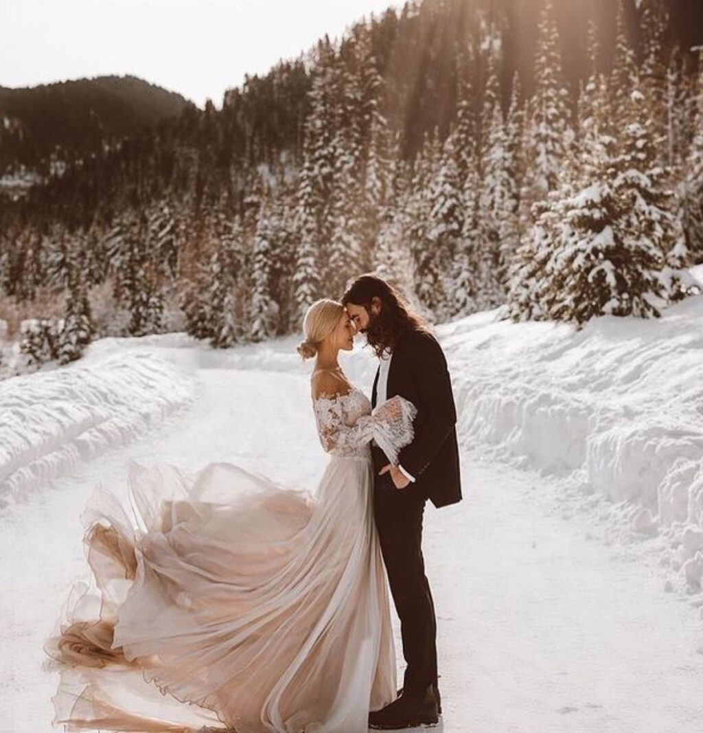 A bride and groom posing in the snow. The bride is wearing a long flowing dress. There are snow covered trees in the background. This is for the blog post called winter wedding picture ideas
