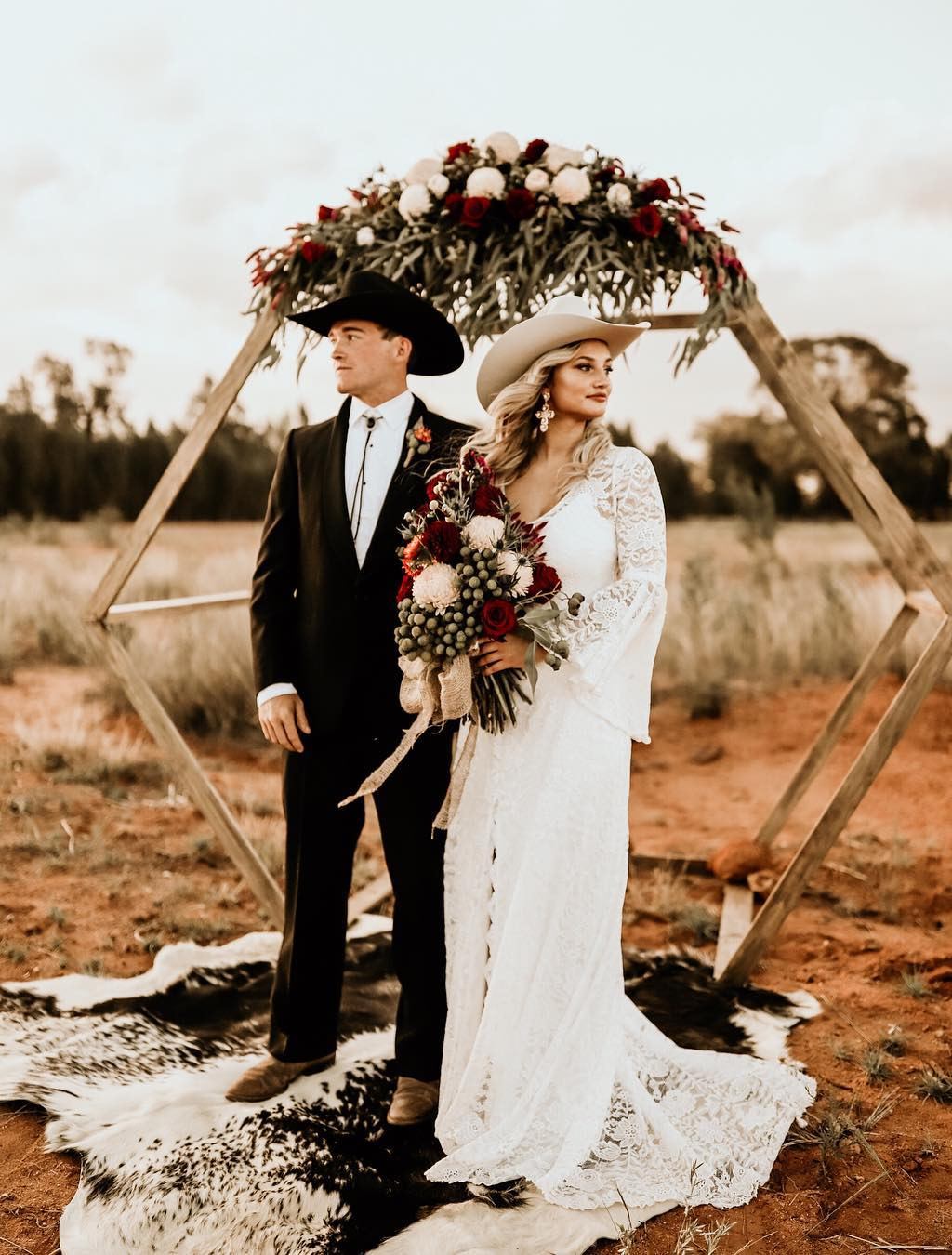 A bride and groom posing in front of a rustic wedding arch with cowboy hats on