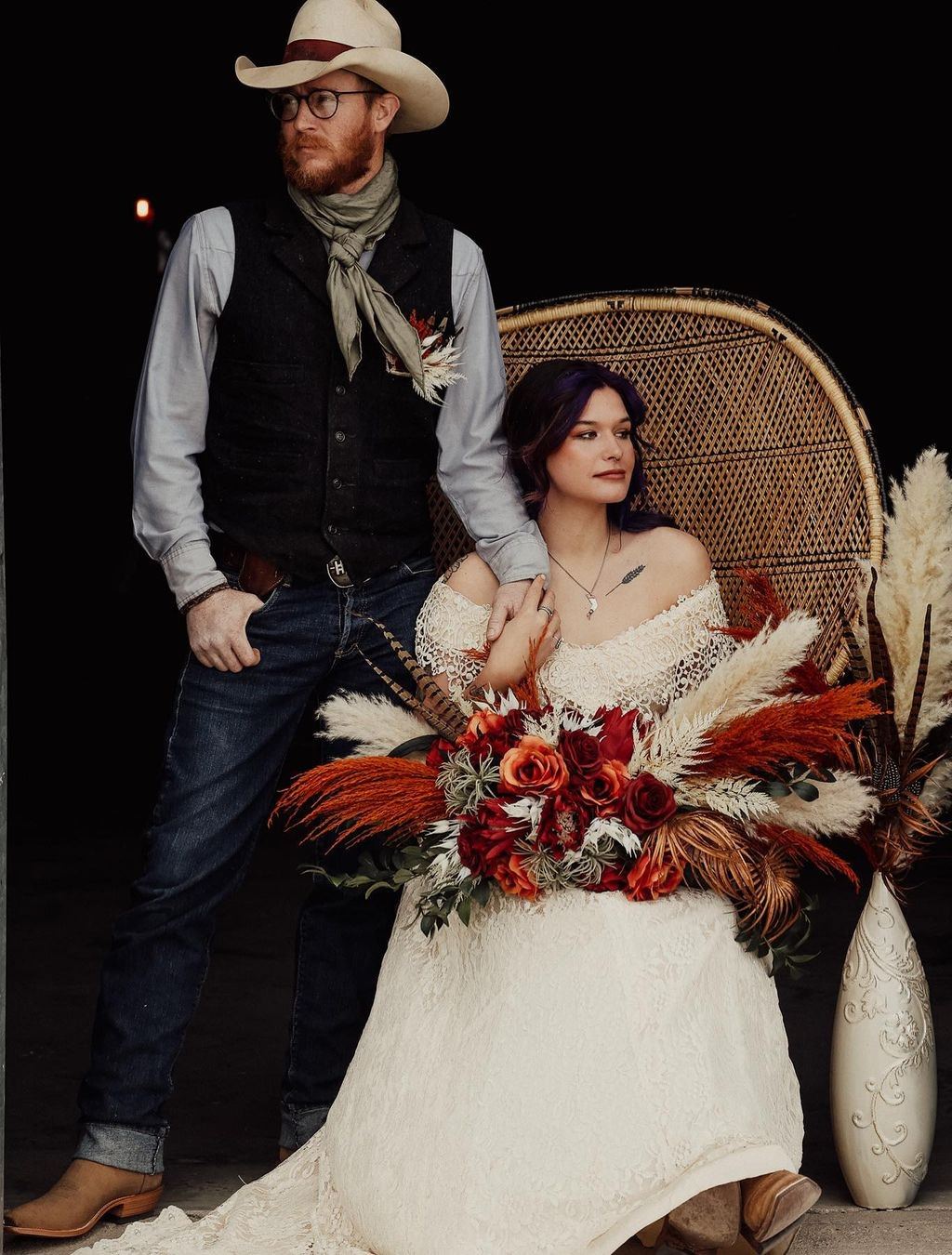 A rustic bride and groom. They are looking in opposite directions. The groom has a cowboy hat and the bride is holding a big boho style bridal bouquet with orange and red glowers and pampas grass
