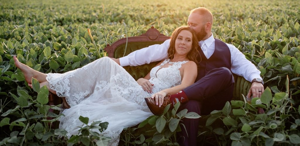 A bride and groom sitting on a couch in the middle of a field. The bride is laying in the groom's lap. She is a barefoot. This is being used for the first image in the blog post called how to plan a wedding in 6 weeks