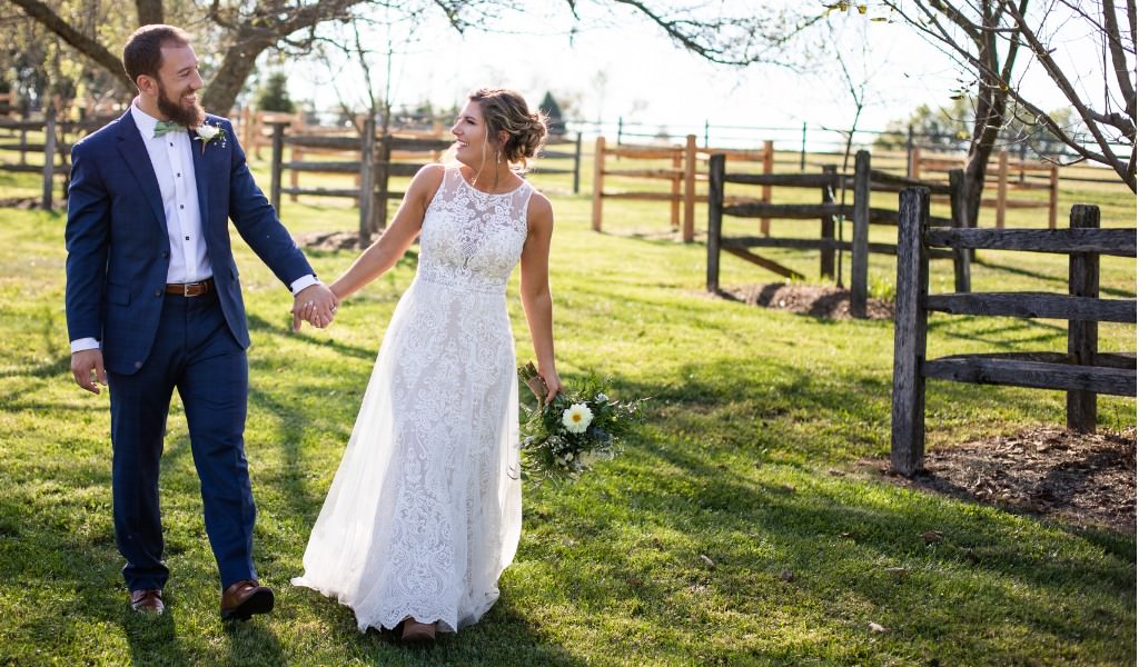 A bride and groom holding hands in a field on their wedding day. They are looking at each other and smiling