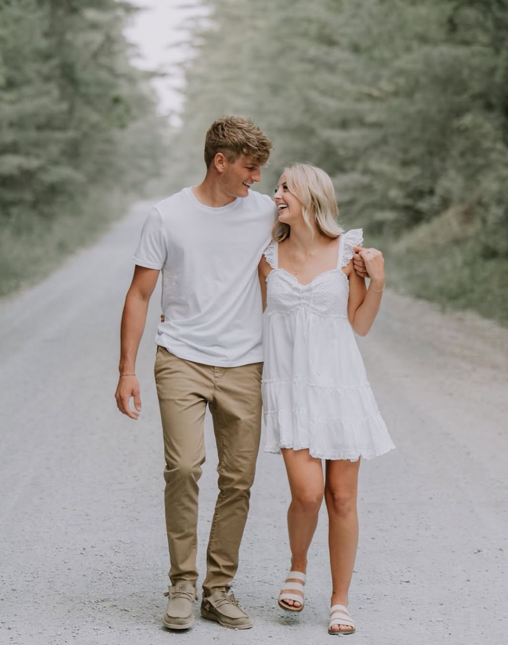 A cute couple walking in the middle of the road for their engagement pictures. They have their arms around each other and are looking at each other and smiling. The girl is in  shot white sundress.