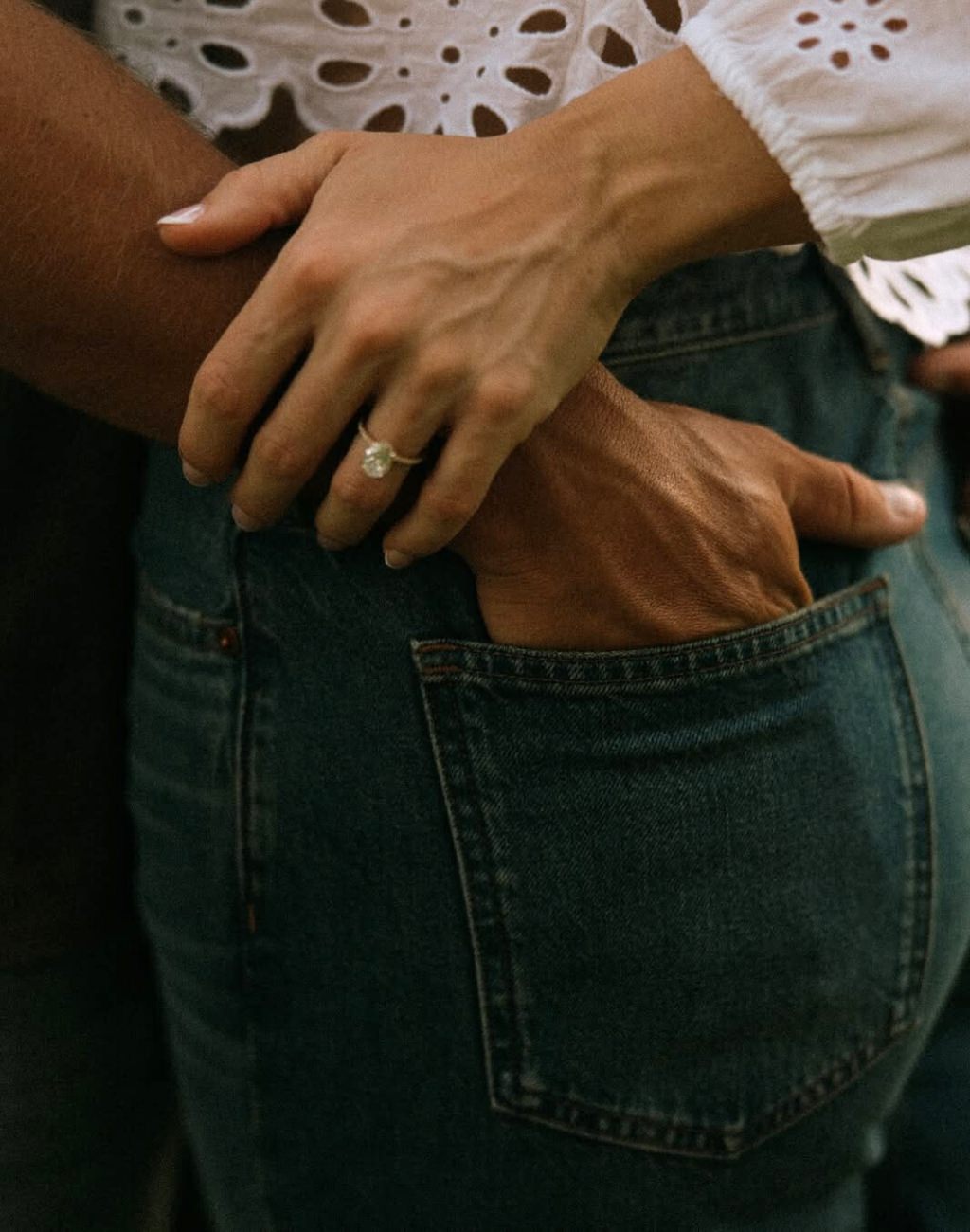 An engagement  photo pose that shows a close up the guy's hand in the girl's back pocket and she is holding his wrist so you can see her engagement ring up close.